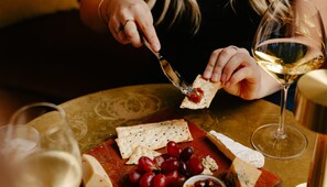 A person holding a cheese knife and a piece of cheese, with a glass of white wine and a plate of cheese and grapes on a table. - Voco Auckland City Centre (Auckland)