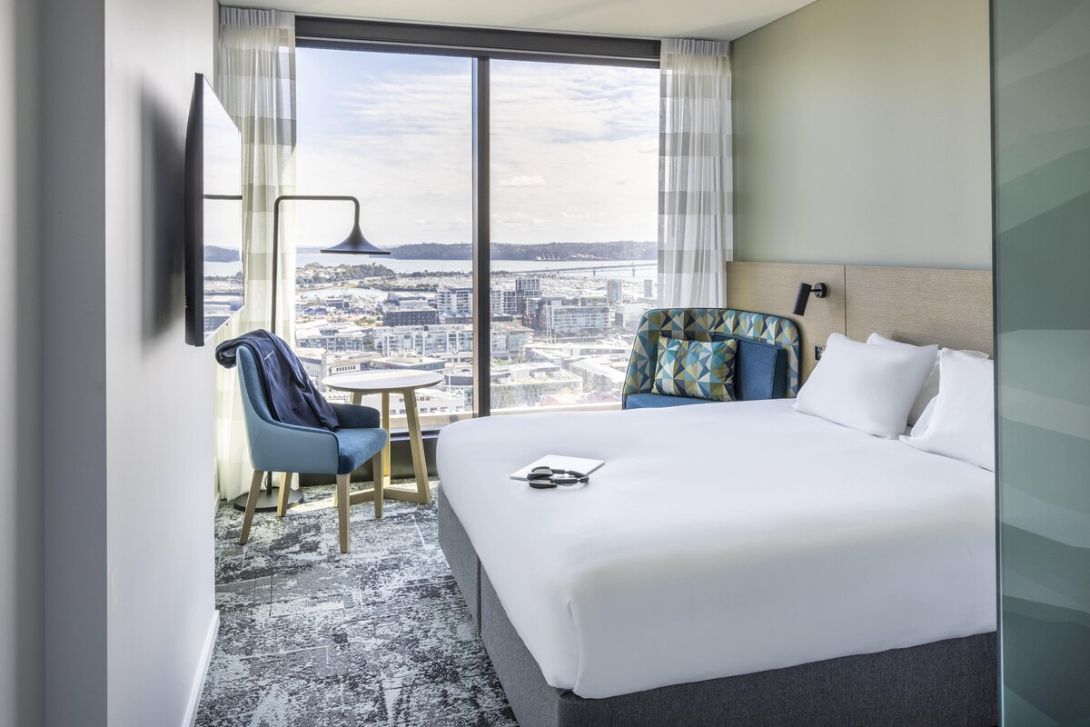 Une chambre d’hôtel avec un grand lit, une chaise, une petite table, un téléviseur et une vue sur le paysage urbain.