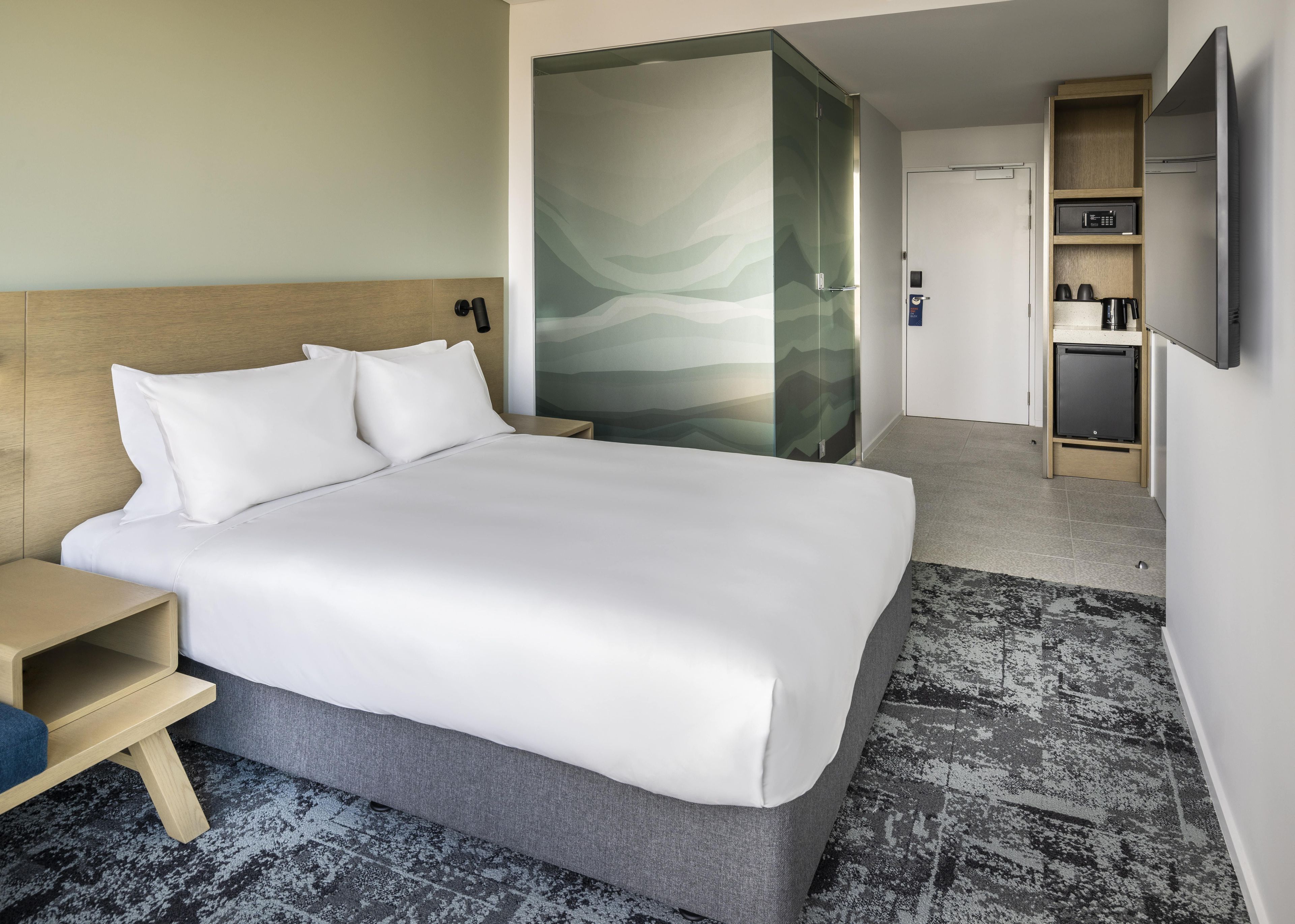 A hotel room with a bed, bedside table, a glass partition, a small kitchenette, and a television.