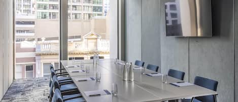 A conference room with a long table, chairs, a flat-screen TV, and large windows overlooking a cityscape.