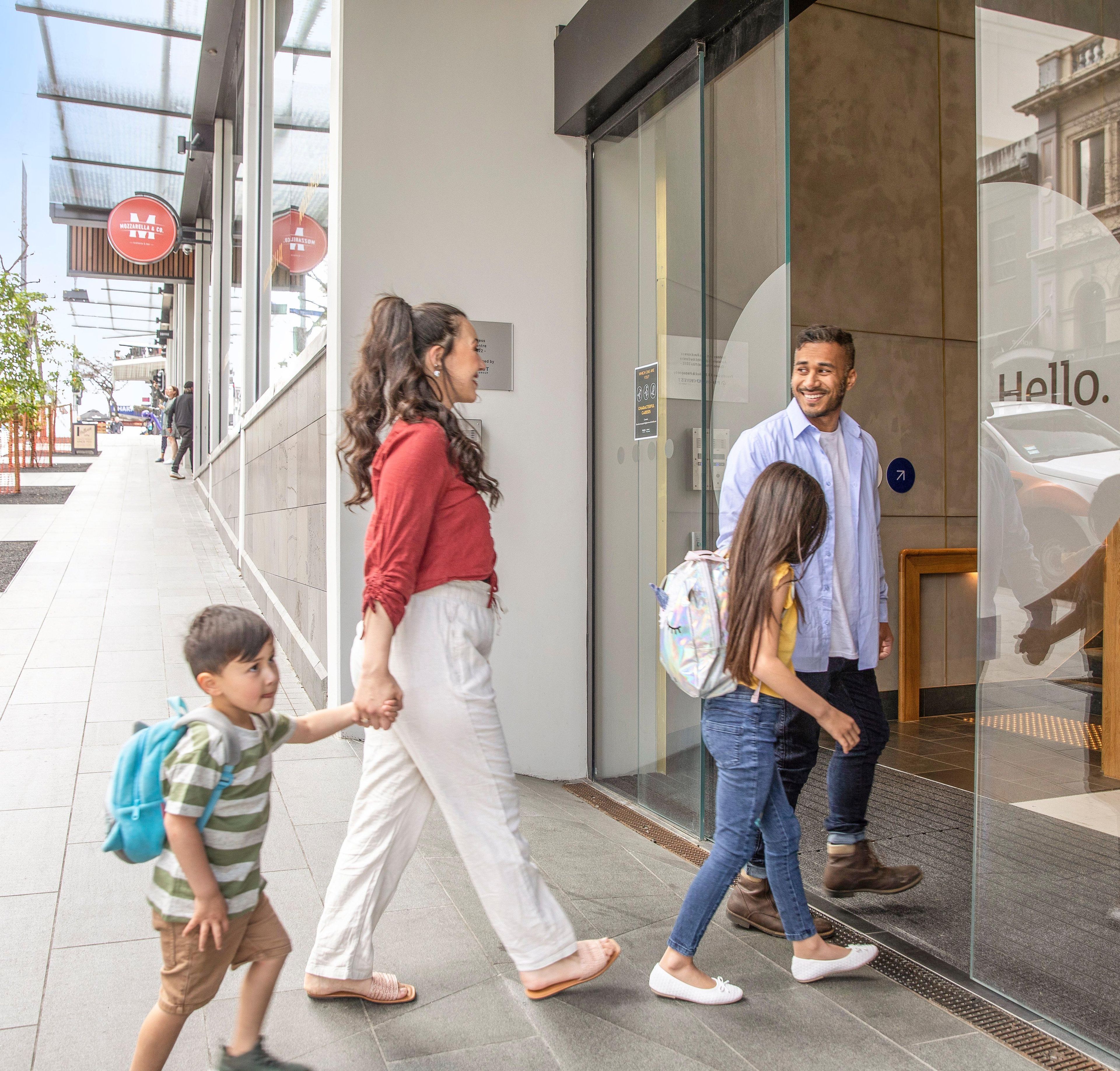 Una familia caminando hacia un edificio con entrada de vidrio y una señal visible.
