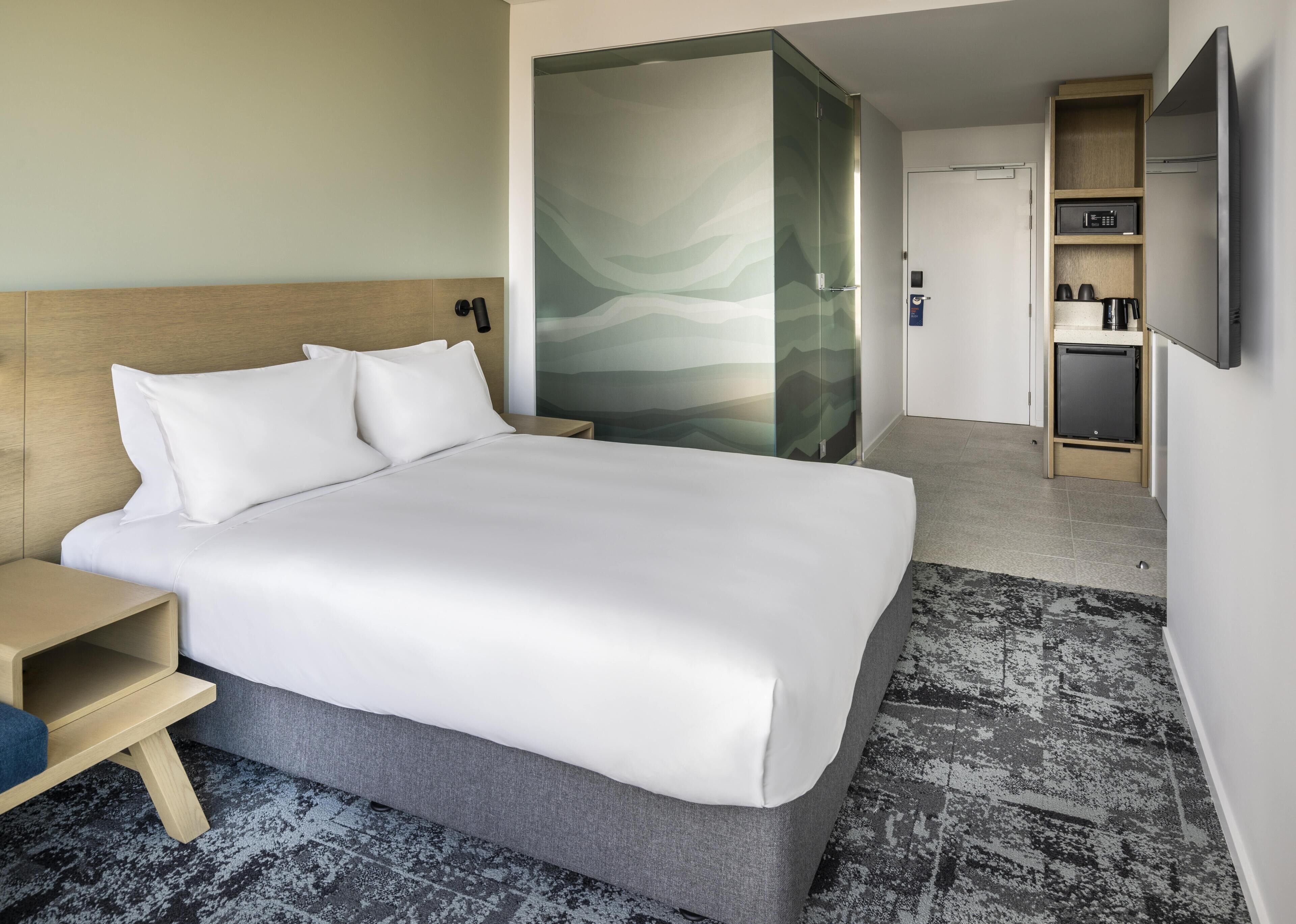 A hotel room with a bed, bedside table, a glass partition, a small kitchenette, and a television.