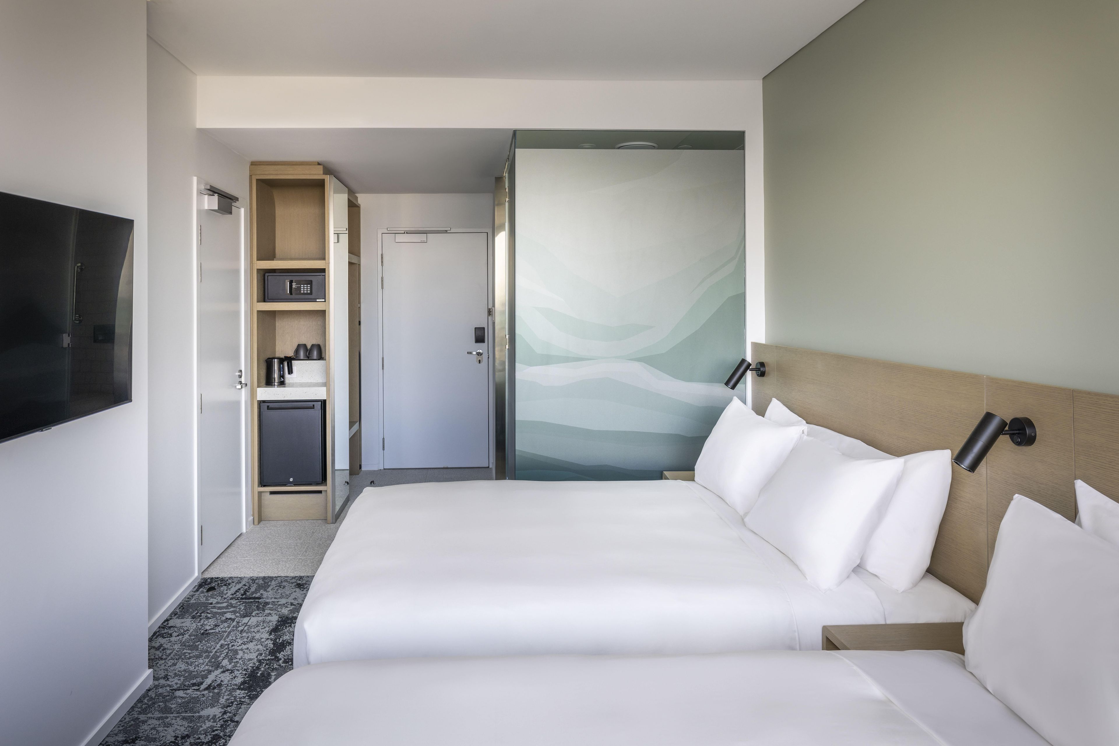 A hotel room with a bed, a TV, a minibar, and a door leading to a bathroom.