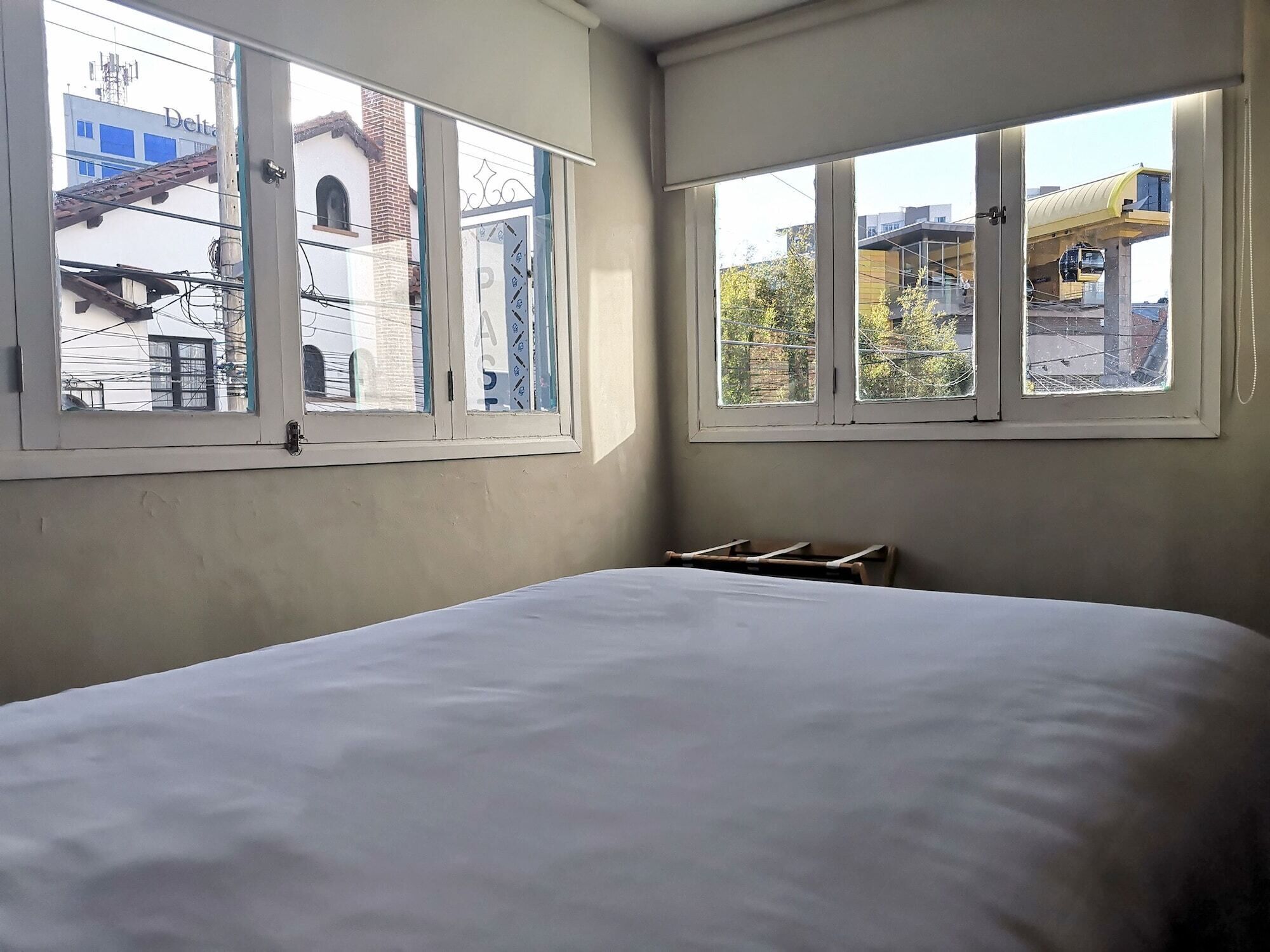 A room with a bed, two windows, and a view of buildings outside.