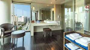 A modern hotel room with a large window, a balcony with wicker furniture, a central bathroom with a mirror, and a glass partition. - Koko Hotel Premier Hakata (Fukuoka)