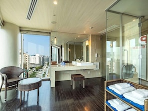 Une chambre d’hôtel moderne dotée d’une grande fenêtre, d’un balcon avec des meubles en osier, d’une salle de bain centrale avec un miroir et d’une cloison en verre.