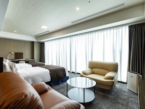 Une chambre d’hôtel avec un lit, un canapé, une chaise, une petite table et une grande fenêtre avec des rideaux.