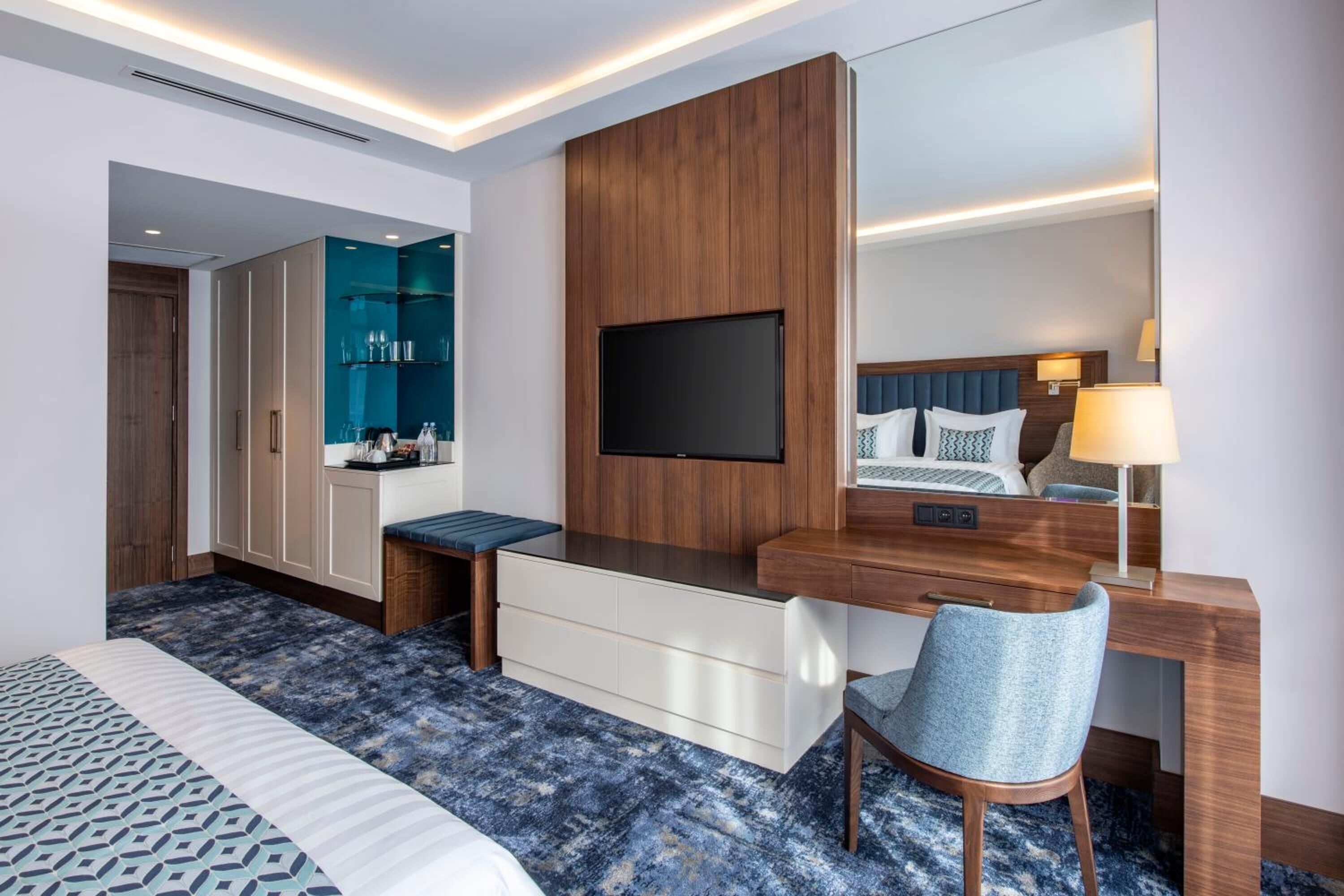 Una habitación de hotel moderna con una cama grande, un escritorio con una silla, un televisor, un espejo y un minibar.