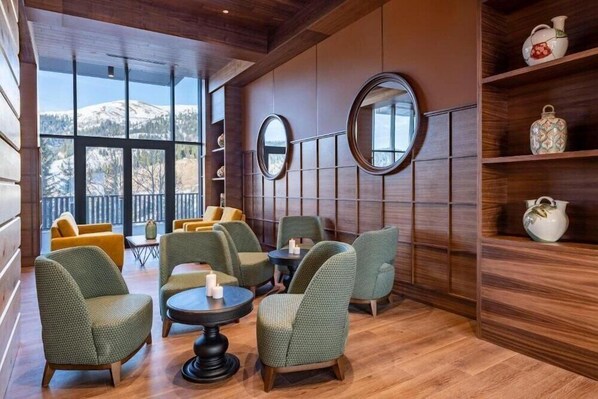 Ein gemütlicher Lounge-Bereich mit grünen Polstersesseln, einem runden Tisch und Holzboden. Große Fenster bieten Blick auf einen verschneiten Berg.