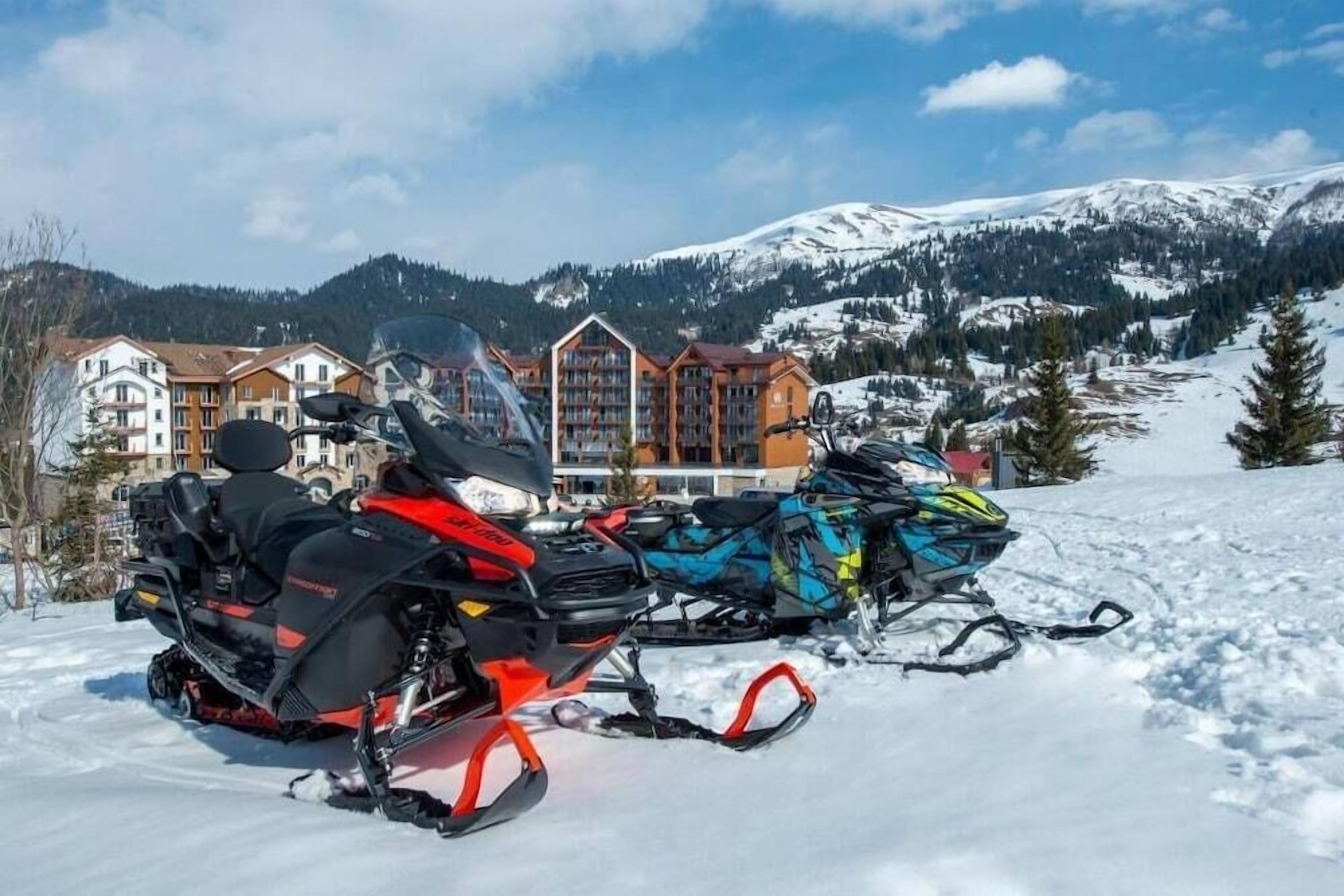 Une motoneige avec un conducteur, un bâtiment doté de nombreux balcons et, au loin, un paysage montagneux enneigé.