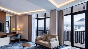 Habitación de hotel con un amplio ventanal que ofrece vistas a la montaña, un sofá, un escritorio y un televisor.