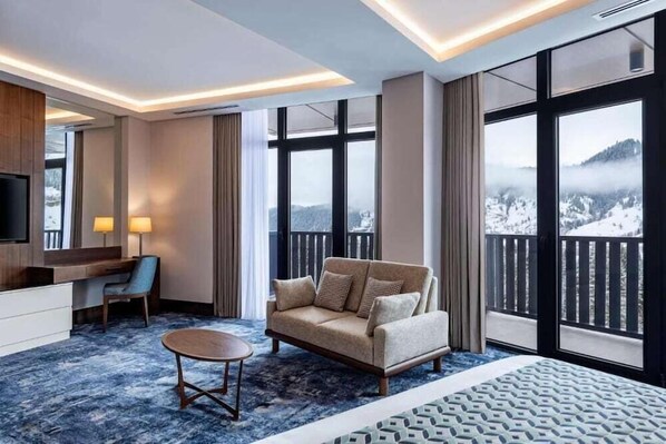 Ein Hotelzimmer mit großem Fenster und Bergblick, einer Couch, einem Schreibtisch und einem Fernseher.
