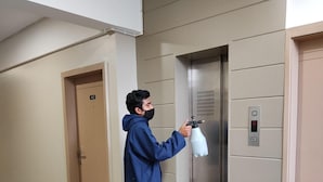 A person in a blue jumpsuit spraying a white object in a hallway with wooden flooring and beige walls.