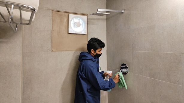 A person cleaning a shower area in a bathroom with tiled walls and a showerhead mounted on the wall. - Kanak Beacon Hotel, Ahmedabad (Ahmedabad)