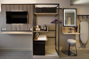 A compact hotel room with a TV, a mirror, a small sink, a stool, and a storage unit with a hanging garment.
