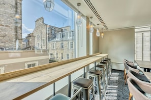 A modern interior with a long wooden counter, bar stools, and large windows offering a view of historic buildings. - Premier Inn Edinburgh City Centre Hotel (Edinburgh)