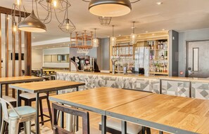 Ein Restaurant mit Holztischen, hängenden Leuchten, einer Bar und einer Küche im Hintergrund.