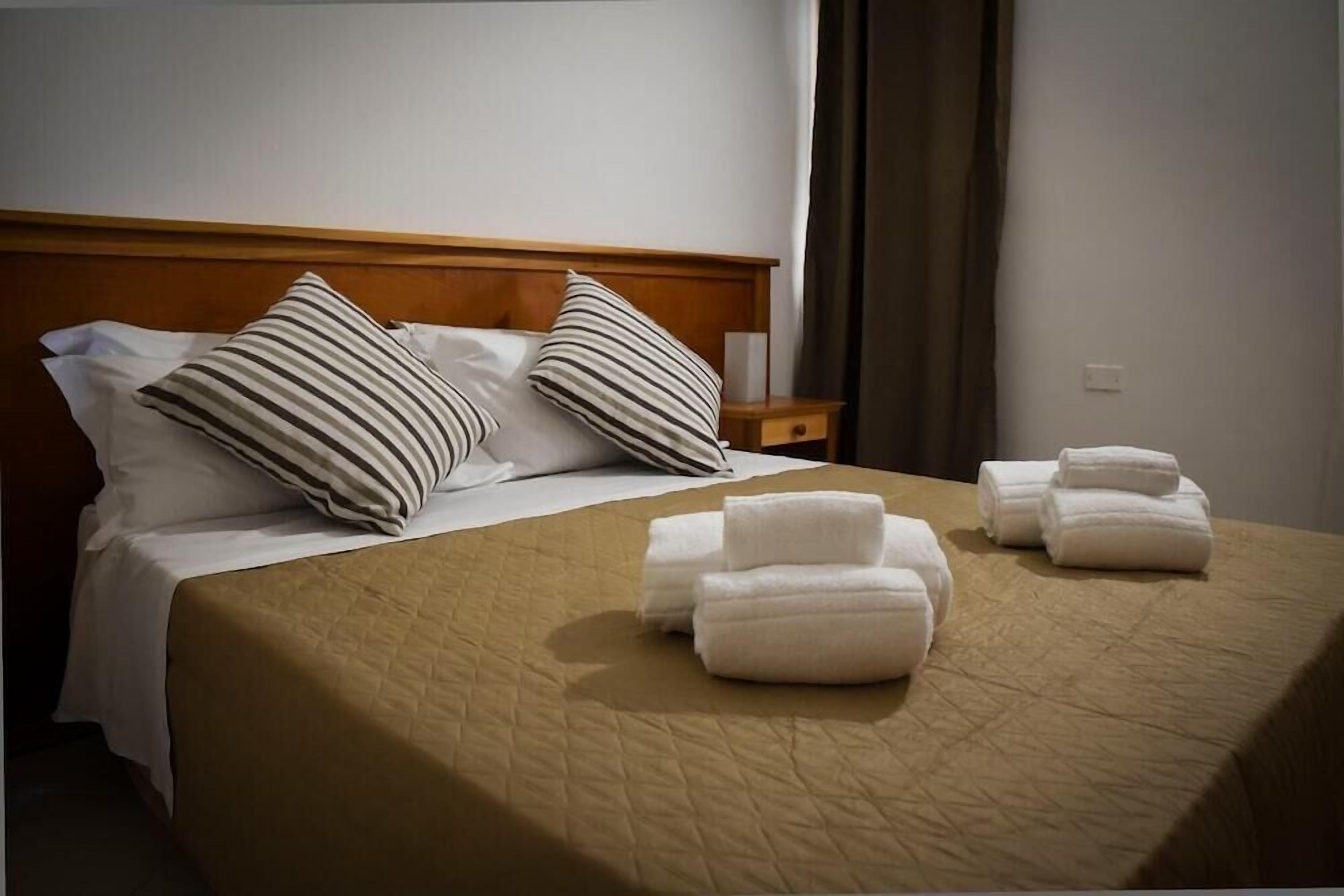 Un letto rifatto con cuscini a righe, una coperta beige e asciugamani piegati su una testiera in legno.