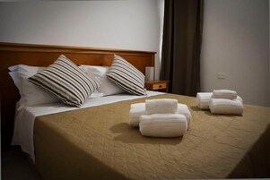 Una cama bien hecha con almohadillas a rayas, una colcha color beige y toallas dobladas sobre un cabecero de madera.