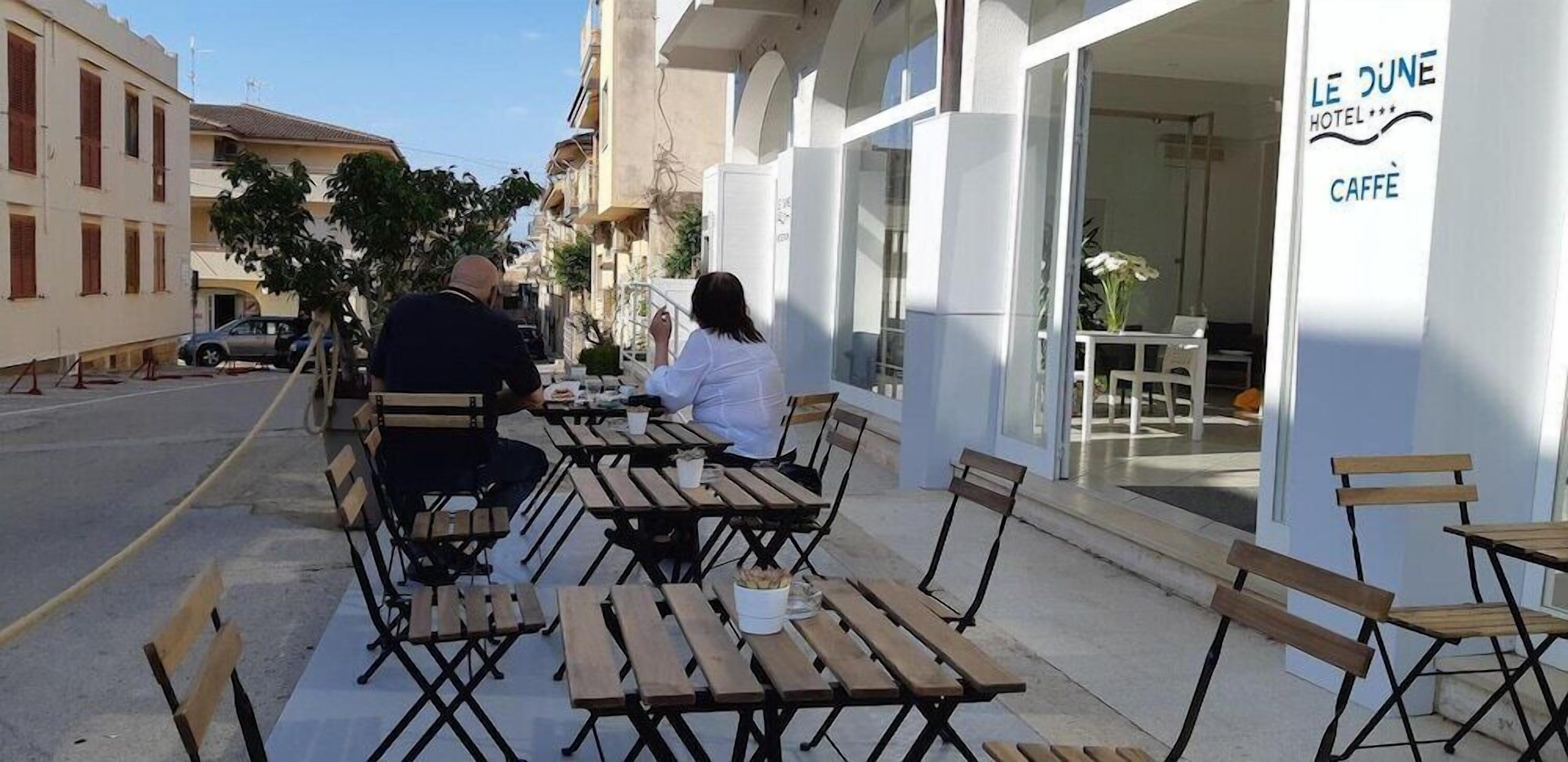 Area esterna con tavoli e sedie di fronte a un edificio con un cartello che riporta la scritta "LE DUNE HOTEL".