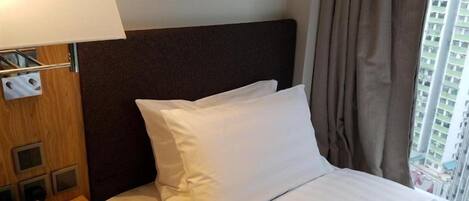 A hotel room with a bed, a bedside table with a lamp, a digital clock, and a window with curtains.