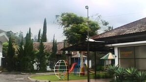 Un parque infantil con toboganes y una estructura para escalar, un área cubierta con un cartel, y un edificio con tejado de baldosas.