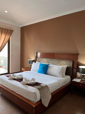 A hotel room with a large bed, a wooden headboard, a nightstand with a lamp, and a window with curtains.