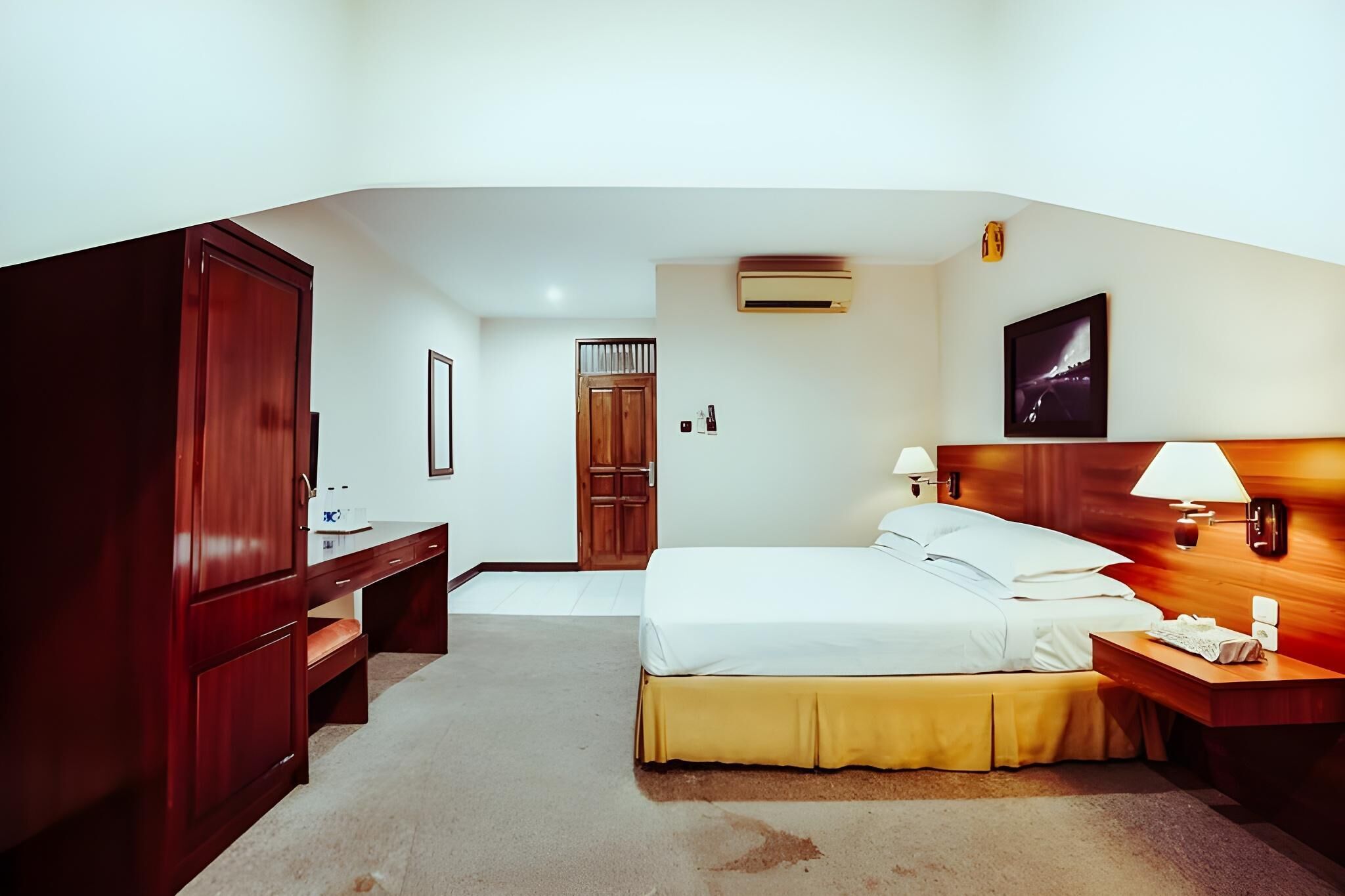 A hotel room with a bed, bedside table, desk, wardrobe, and an air conditioning unit.