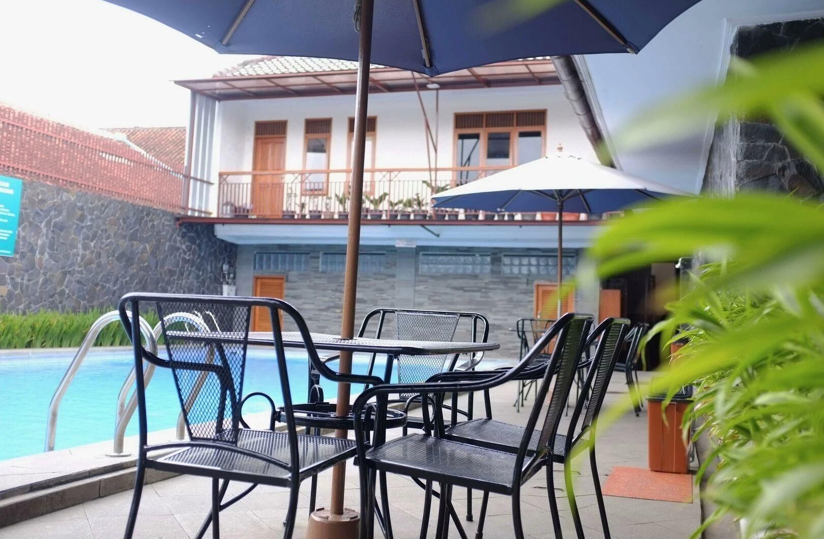 A poolside area with metal chairs and tables, a stone wall, and a building with a balcony in the background.
