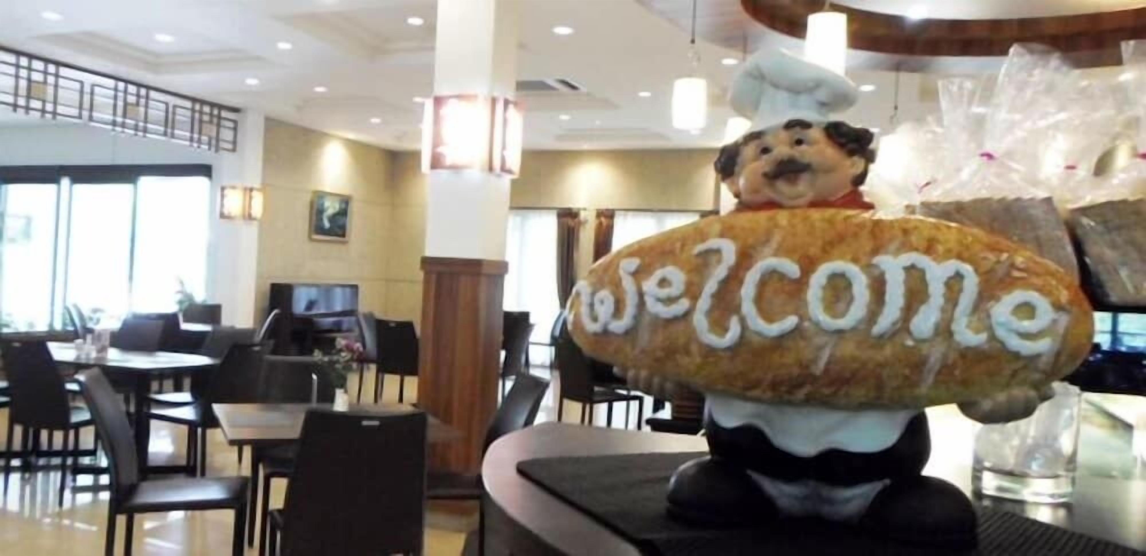 En restaurantindretning med borde og stole, en stor stuffedyr med 'Welcome' skrevet på, og en kokkefigur.