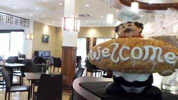 En restaurantindretning med borde og stole, en stor stuffedyr med 'Welcome' skrevet på, og en kokkefigur.