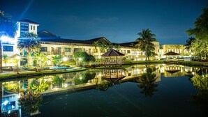 A nighttime view of a hotel with a reflective pond, palm trees, and a gazebo. - Rukun Resort Sentul (Citaringgul)
