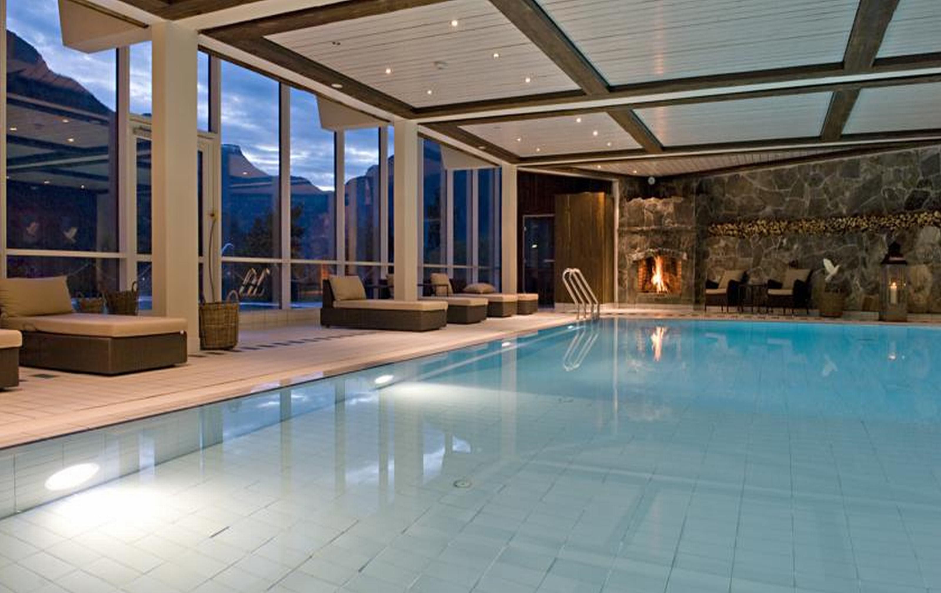Piscina interior moderna com parede de pedra e lareira, grandes janelas e área de estar externa.
