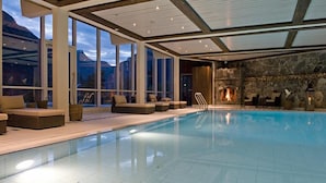 A modern indoor swimming pool with a stone wall and a fireplace, large windows, and outdoor seating. - Hotel Union Geiranger (Stranda)