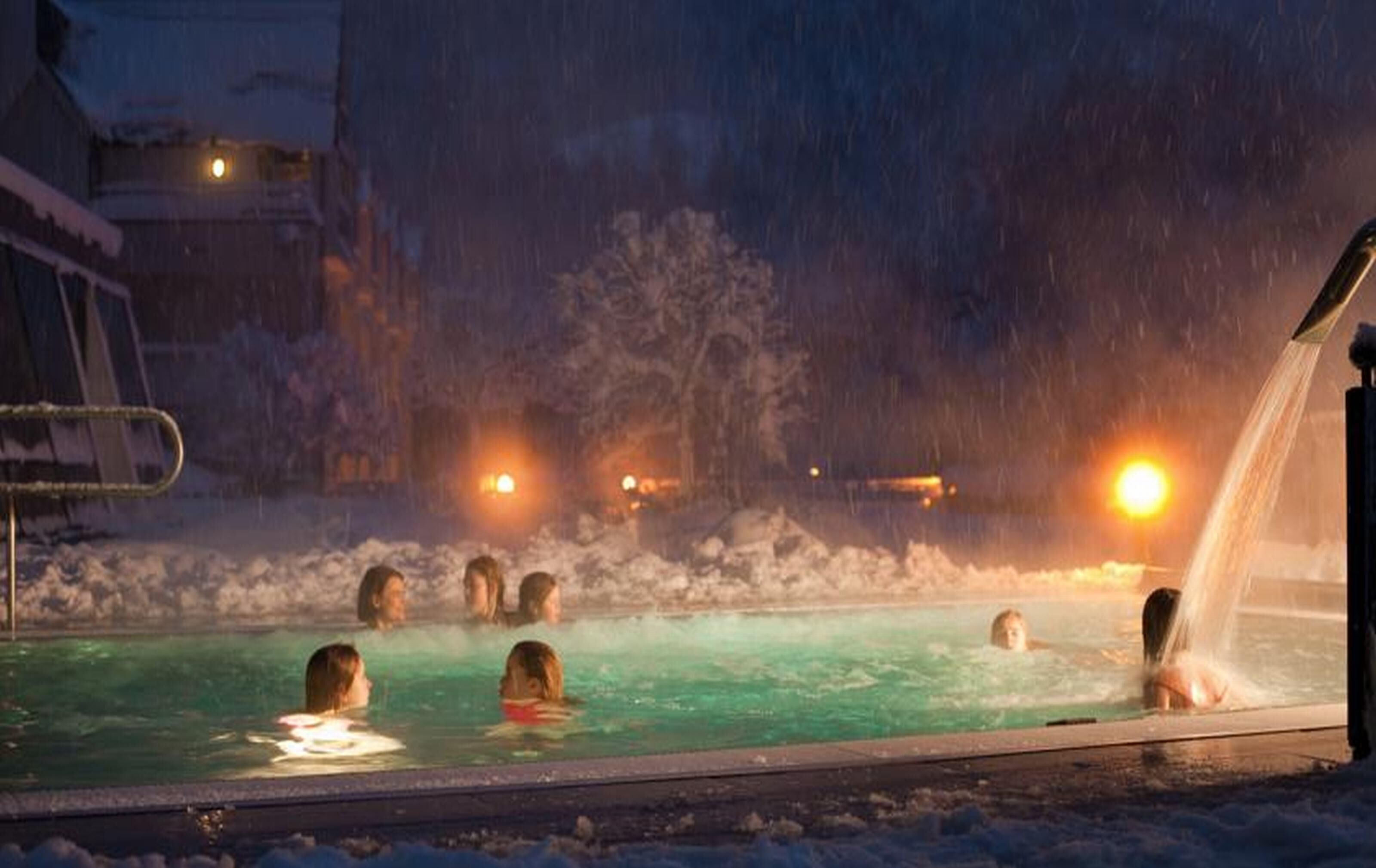 Zona de piscina exterior cubierta de nieve, con gente nadando y un jacuzzi a la derecha.