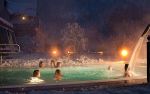 A snow-covered outdoor pool area with people swimming and a hot tub on the right.