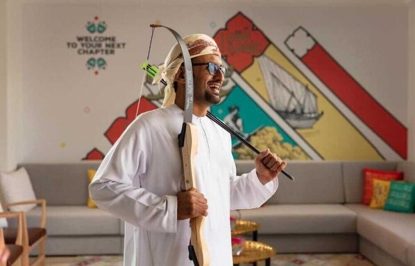 A person in traditional attire holding a bow and arrow, standing in a room with a colorful wall mural and a sofa. - Atana Stay Al Ashkhara (Al Ashkharah)