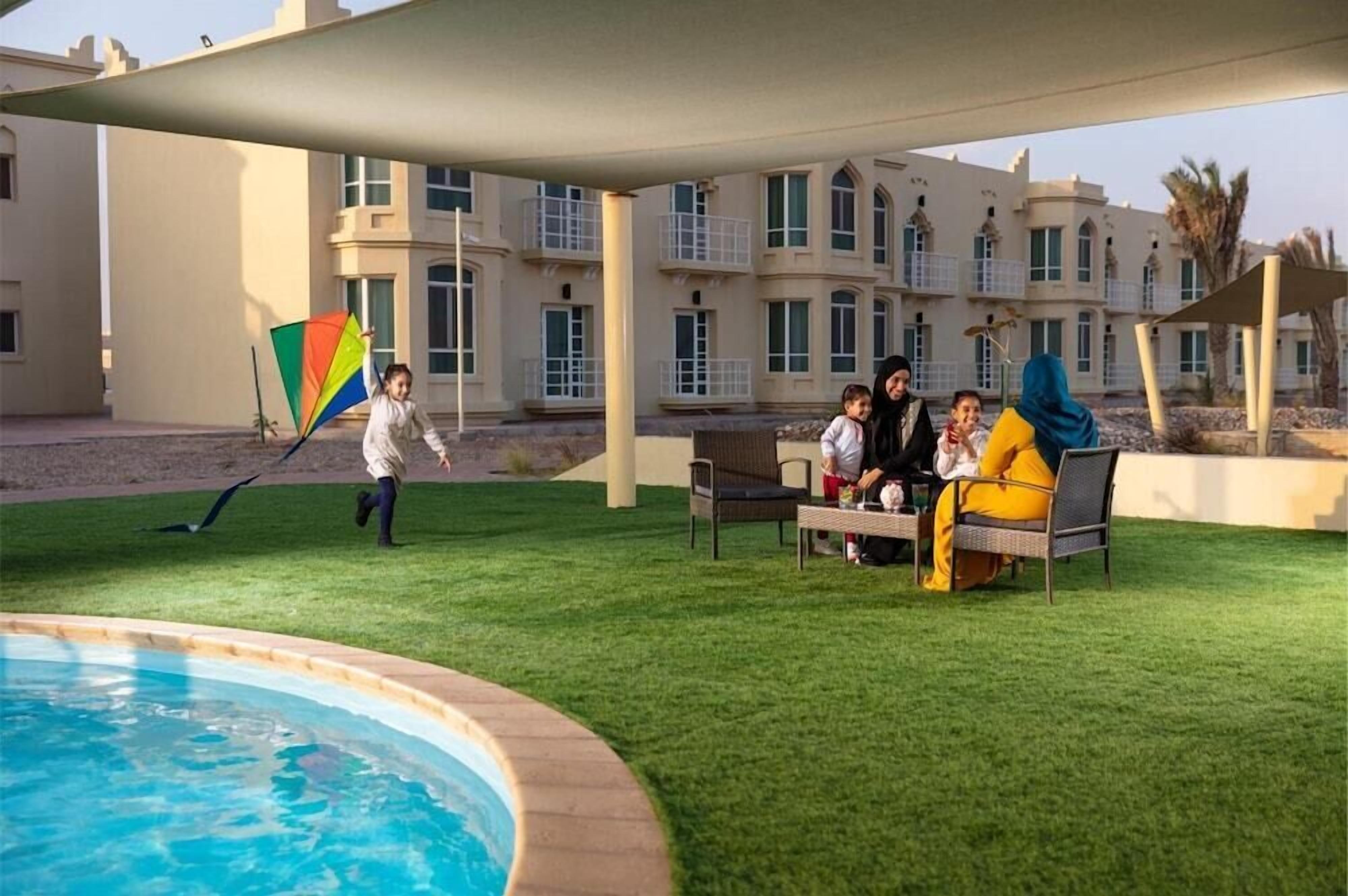 A poolside area with a grassy lawn, lounge chairs, and a building with multiple windows and balconies in the background.