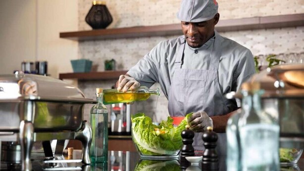 Un chef prepara comida en una cocina con pared de ladrillo, estantes y diversos electrodomésticos.