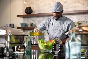 A chef preparing food in a kitchen with a brick wall, shelves, and various kitchen appliances. - Atana Stay Al Ashkhara (Al Ashkharah)