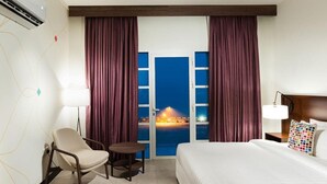 Habitación de hotel con cama, una silla, una mesita, una lámpara y vista a la playa de noche.