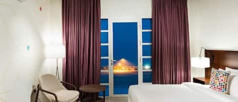 A hotel room with a bed, a chair, a small table, a lamp, and a view of a beach at night.