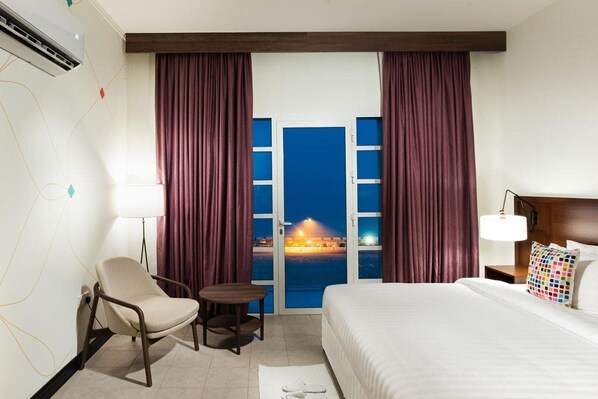 A hotel room with a bed, a chair, a small table, a lamp, and a view of a beach at night. - Atana Stay Al Ashkhara (Al Ashkharah)