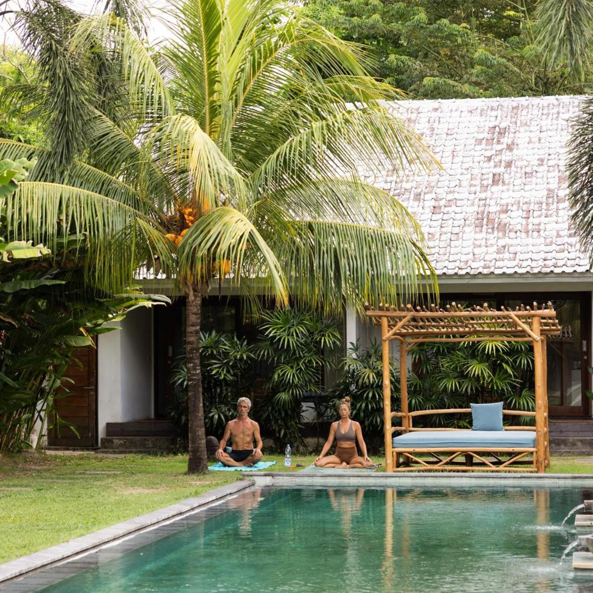 Une piscine où deux personnes méditent, entourées d’une végétation luxuriante et surmontée d’un toit traditionnel recouvert de tuiles.