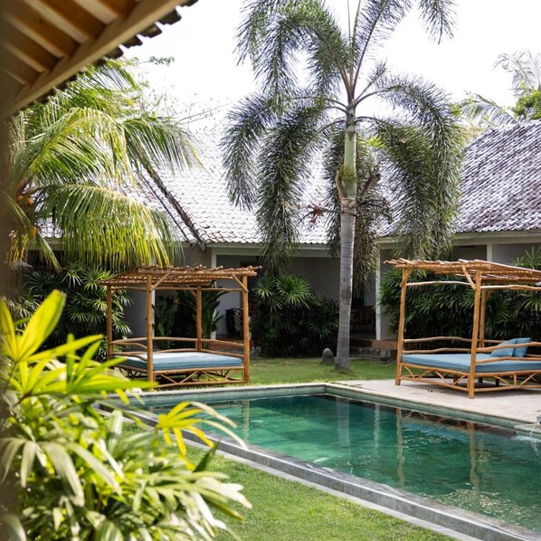 A pool area with two wooden loungers, a palm tree, and a tiled roof. - Kuta Baru Hotel (Kuta)