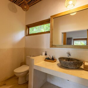 A bathroom with a white toilet, a stone sink, a wooden-framed mirror, and a window with a view of greenery. - Kuta Baru Hotel (Kuta)