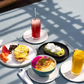Una mesa con diversos alimentos y bebidas, incluyendo un batido, un plato de comida con un topping colorido, y una tostada con huevo poché y aguacate.