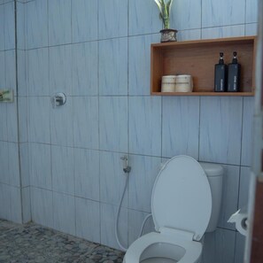 A bathroom with a white toilet, a shower area, and a shelf with toiletries. - Pakels Bali Villas (Karangasem)