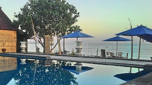 A pool with clear water reflecting trees and a view of the sea. There are lounge chairs and umbrellas along the poolside. - Pakels Bali Villas (Karangasem)