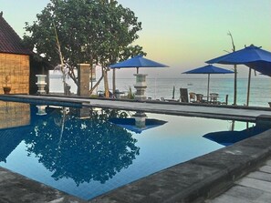 A pool with clear water reflecting trees and a view of the sea. There are lounge chairs and umbrellas along the poolside. - Pakels Bali Villas (Karangasem)
